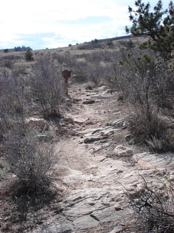 A rocky trail winding through a sparse landscape of dry shrubs and grass, with a wooden signpost visible in the background. The sky is partly cloudy, and gently rolling hills can be seen in the distance. Rabbit Mountain Loop mountain bike trail.