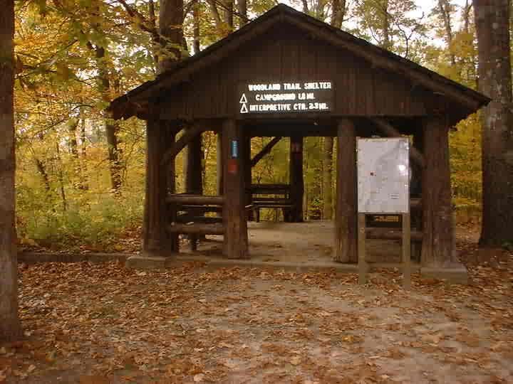 A rustic wooden trail shelter surrounded by autumn foliage. The shelter has a sign reading "Woodland Trail Shelter" along with information about camping and nearby interpretive trails. The ground is covered with fallen leaves, and picnic tables can be seen inside the structure. Bluff Trail mountain bike trail.
