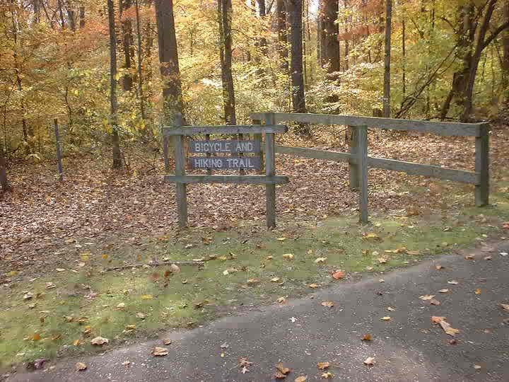 A wooden sign marking the entrance to a bicycle and hiking trail, surrounded by vibrant autumn foliage and fallen leaves on the ground. A gravel path leads into the wooded area, indicating a nature trail suitable for outdoor activities. Bluff Trail mountain bike trail.