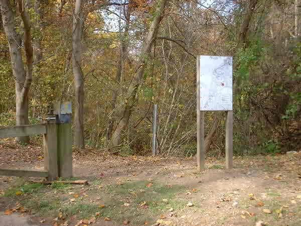 A pathway leading into a wooded area, marked by a wooden gate with a latch. Beside the path, a white informational sign stands on two posts, surrounded by trees with autumn foliage. The ground is covered with fallen leaves. Bluff Trail mountain bike trail.