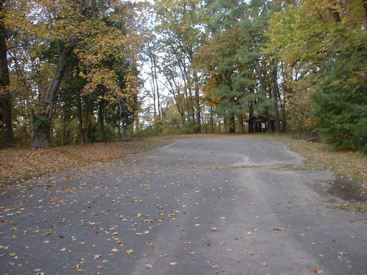 A paved pathway surrounded by trees, covered with fallen autumn leaves. In the background, a small structure can be seen partially obscured by foliage, suggesting a tranquil outdoor setting. Bluff Trail mountain bike trail.