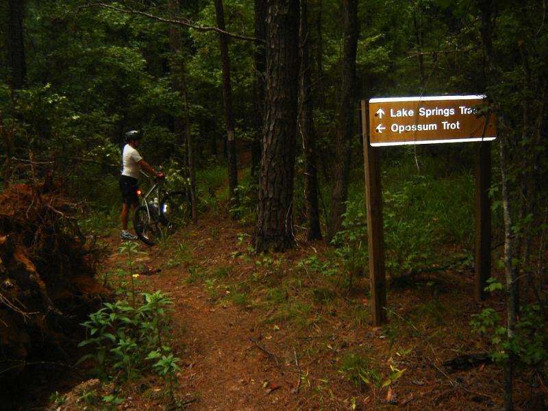 A mountain biker stands next to a sign directing to Lake Springs Trail and Opossum Trot in a wooded area. The path is surrounded by tall trees and lush greenery, with a fallen log visible on the ground. Bartram Trail / West Dam / Wildwood Park mountain bike trail.