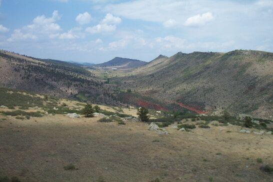 A scenic view of rolling hills and valleys under a partly cloudy sky, with grassy terrain and scattered trees. The landscape features a winding path or road visible in the distance, leading through the natural setting. Rabbit Mountain Loop mountain bike trail.
