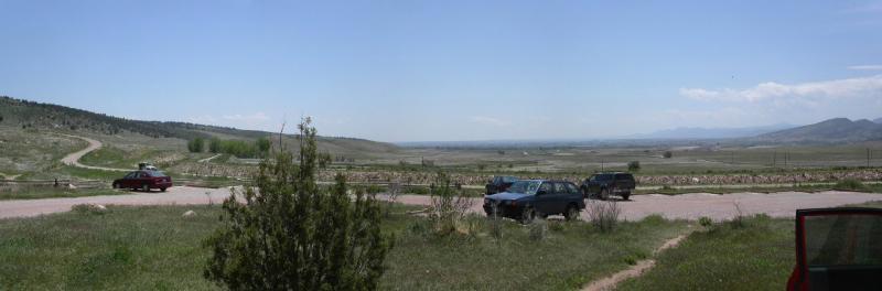 A panoramic view of a scenic landscape featuring rolling hills under a clear blue sky. In the foreground, two parked cars are visible on a gravel road, surrounded by lush greenery and sparse vegetation. The background showcases expansive fields leading towards distant mountains. Rabbit Mountain Loop mountain bike trail.
