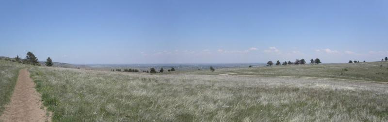Panoramic view of a grassy landscape with a dirt path running through the foreground, leading to rolling hills under a clear blue sky with scattered clouds. Rabbit Mountain Loop mountain bike trail.