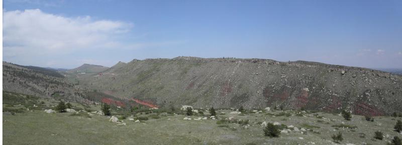 Panoramic view of a rugged landscape, featuring rolling hills and rocky terrain under a bright blue sky with scattered clouds. The foreground includes patches of green grass and small trees, while the background showcases a prominent ridge with textured rock formations. Rabbit Mountain Loop mountain bike trail.