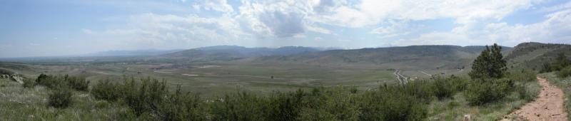 A panoramic view of a vast landscape featuring rolling hills, scattered shrubs, and a winding path leading into the distance. The sky is partly cloudy, with patches of blue and soft white clouds. The scene conveys a sense of openness and natural beauty. Rabbit Mountain Loop mountain bike trail.