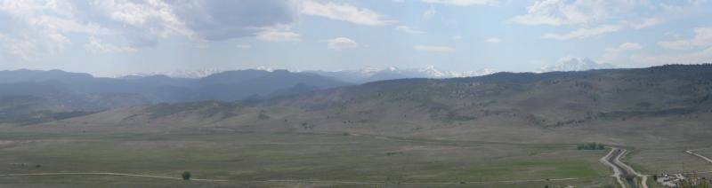 Panoramic view of rolling hills and mountains under a partly cloudy sky, with a winding road visible in the foreground. The landscape features open fields and distant snow-capped peaks. Rabbit Mountain Loop mountain bike trail.