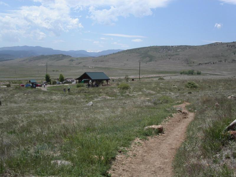 A scenic view of a grassy landscape with a winding dirt path leading towards a pavilion with a green roof. In the background, rolling hills and mountains can be seen under a partly cloudy sky. A few parked cars and people are near the pavilion, surrounded by open fields. Rabbit Mountain Loop mountain bike trail.