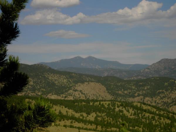 A scenic view of rolling mountain ranges under a partly cloudy sky, with a foreground of trees and varying shades of green hills. The distant mountains feature peaks that rise prominently against the horizon. Heil Valley Ranch mountain bike trail.