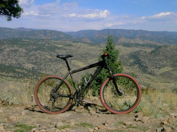 A mountain bike positioned on rocky ground, overlooking a scenic valley with rolling hills and distant mountains under a partly cloudy sky. The bike features black and red tires and is surrounded by tall grass and sparse vegetation. Heil Valley Ranch mountain bike trail.
