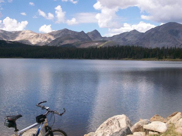 A serene lakeside scene featuring a calm body of water reflecting a clear blue sky with fluffy white clouds. In the foreground, a mountain bike is resting on large rocks at the water's edge, surrounded by lush greenery and distant mountains. The landscape conveys a sense of tranquility and the beauty of nature. Sourdough Trail mountain bike trail.