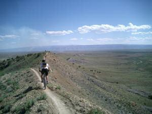 A person riding a mountain bike along a narrow trail on a ridge, with a panoramic view of rolling hills and a clear blue sky in the background. Zippety Do Dah mountain bike trail.