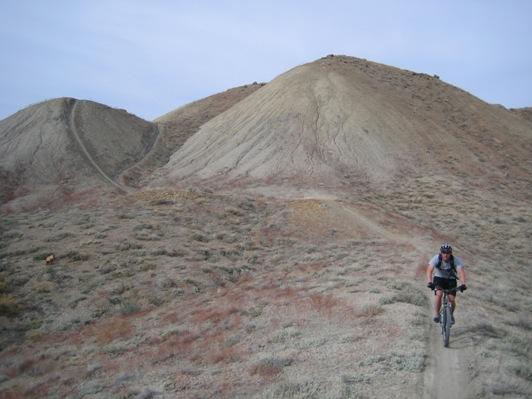 A mountain biker riding on a dirt trail through a hilly, arid landscape with sparse vegetation. The background features rolling hills with a gentle slope, under a clear sky. Zippety Do Dah mountain bike trail.