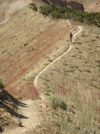 A winding dirt path leads through a hilly landscape with sparse vegetation, where a person is seen biking along the trail. The terrain features a mix of brown and green, and the surrounding area appears arid and hilly. Zippety Do Dah mountain bike trail.