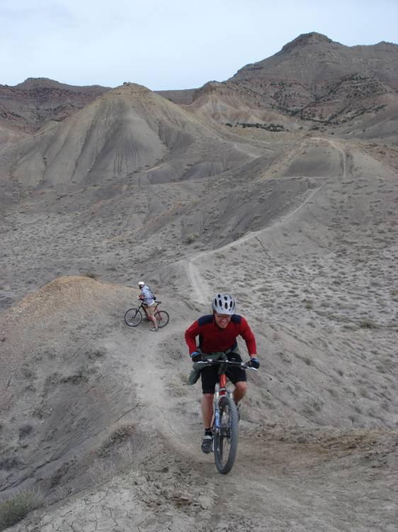 Two mountain bikers navigate a rugged, arid landscape with hills and dirt paths. One rider, wearing a red long-sleeve shirt and helmet, is climbing up a steep incline, while the other, dressed in a white jersey and shorts, rides on a lower trail. The terrain is dry and dusty, showcasing the natural contours of the mountains in the background under a cloudy sky. Zippety Do Dah mountain bike trail.