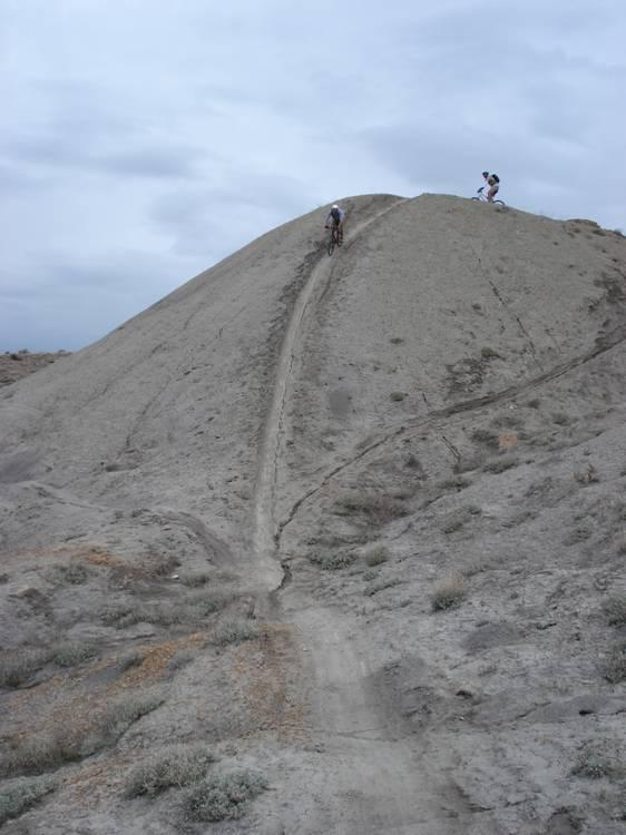 Two mountain bikers navigating a steep, dirt trail on a barren hill, surrounded by sparse vegetation under a cloudy sky. Zippety Do Dah mountain bike trail.