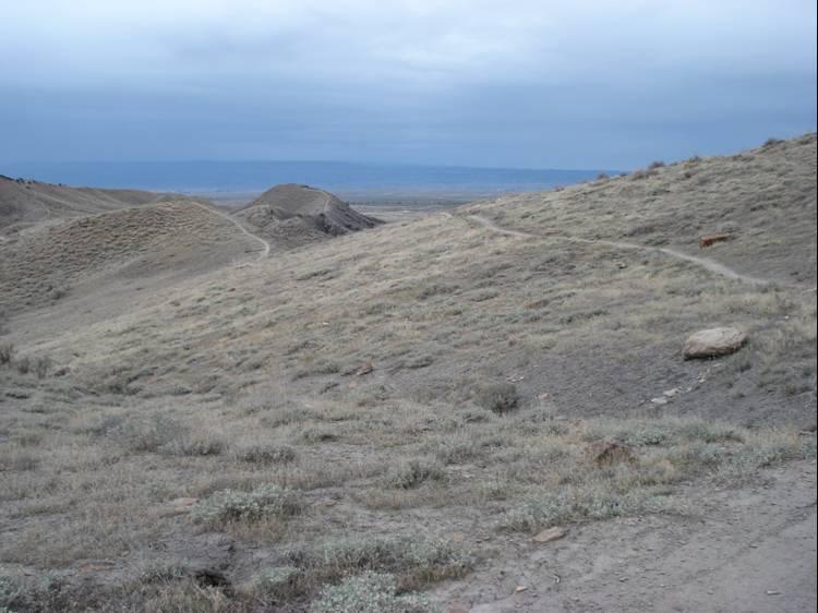 A scenic view of rolling hills covered in dry grass and scattered rocks, set under a cloudy sky. The landscape stretches into the distance, with a winding path visible among the hills. Zippety Do Dah mountain bike trail.