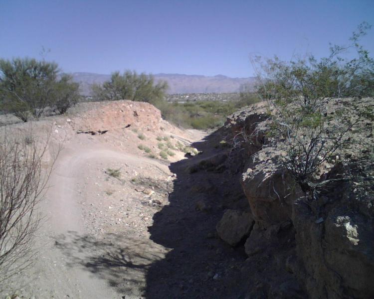 A dirt trail winding between rocky hills in a semi-arid landscape under a clear blue sky, with sparse vegetation and distant mountains visible in the background. Fantasy Island mountain bike trail.