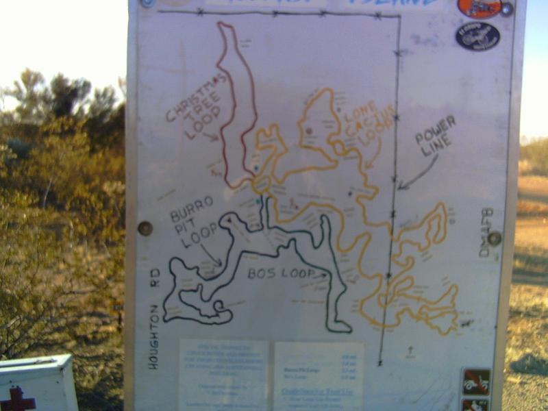 Map of a trail system showing various loop trails, including Christmas Tree Loop, Burro Pit Loop, and Bos Loop, highlighted in different colors. The map is mounted on a post in a desert landscape, with labels indicating roads and nearby features. Fantasy Island mountain bike trail.