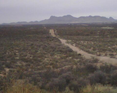 A dusty road winding through a sparse desert landscape, leading towards distant mountains under a cloudy sky. The foreground features low shrubs and grasses typical of a desert environment. Huachuca Canyon mountain bike trail.
