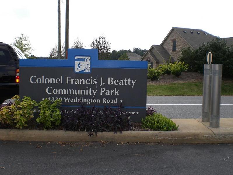 Sign for Colonel Francis J. Beatty Community Park, located at 43230 Weddington Road, featuring the park’s name, a walking figure icon, and surrounding landscaping with greenery. Colonel Francis Beatty Park mountain bike trail.