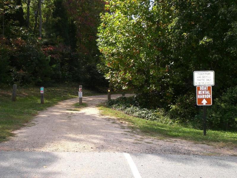 Alt text: A dirt path leading into a wooded area, flanked by green trees and foliage. Signs are posted along the trail, including one indicating "Boat Rental Harbor" with an arrow pointing in the direction of the rental location. Colonel Francis Beatty Park mountain bike trail.