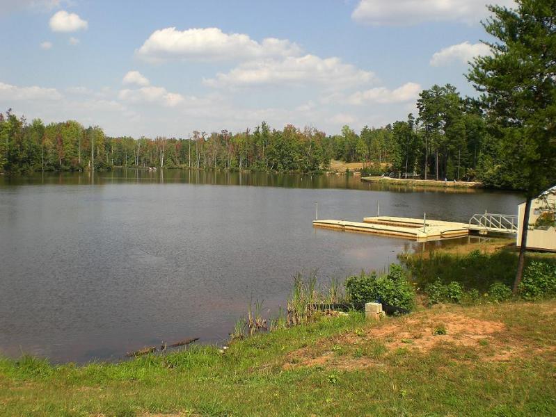 A peaceful lake scene surrounded by trees, with a partially cloudy blue sky. The lake reflects the greenery of the forest, and two wooden docks are visible at the water's edge. Lush grass and shoreline add to the natural beauty of the landscape. Colonel Francis Beatty Park mountain bike trail.