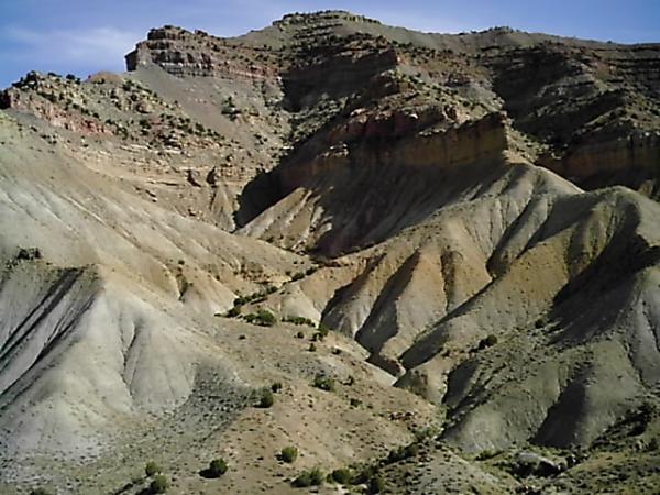A scenic view of a rugged, multi-colored mountain landscape featuring steep, rolling hills and layered rock formations under a clear blue sky. Sparse vegetation dots the area, adding a touch of greenery to the arid terrain. Zippety Do Dah mountain bike trail.