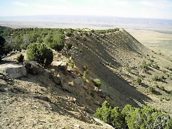 A panoramic view of a rugged landscape featuring a steep, sloping hillside covered with sparse vegetation and scattered rocks. In the background, expansive plains stretch toward the horizon, under a partly cloudy sky. Zippety Do Dah mountain bike trail.