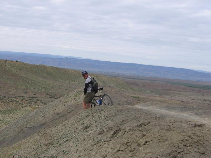 A mountain biker standing on a ridge, looking back while holding a bicycle. The landscape features rolling hills and a vast, open sky overhead, creating a scenic view of the terrain. Zippety Do Dah mountain bike trail.