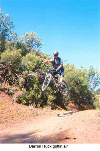A mountain biker performing a jump over a dirt path, surrounded by shrubs and trees under a clear blue sky. The rider is wearing a helmet and casual biking attire, showcasing a moment of athleticism and skill. Downieville Downhill mountain bike trail.