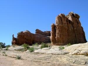 A sunlit rocky formation with towering reddish-brown cliffs set against a clear blue sky. The foreground features a sandy surface with sparse vegetation. Monitor &amp; Merrimac/Courthouse Pasture mountain bike trail.