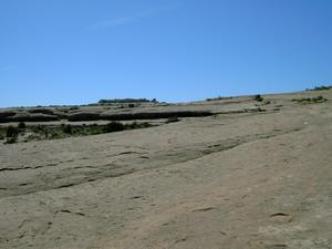 A vast, rocky landscape with a smooth, sloping surface under a clear blue sky. Sparse vegetation is visible in the foreground, and the horizon features rolling terrain in the background. Monitor &amp; Merrimac/Courthouse Pasture mountain bike trail.