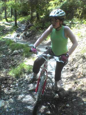 A person wearing a helmet and a green tank top is standing next to a mountain bike on a rocky trail surrounded by forest. Kirroughtree mountain bike trail.