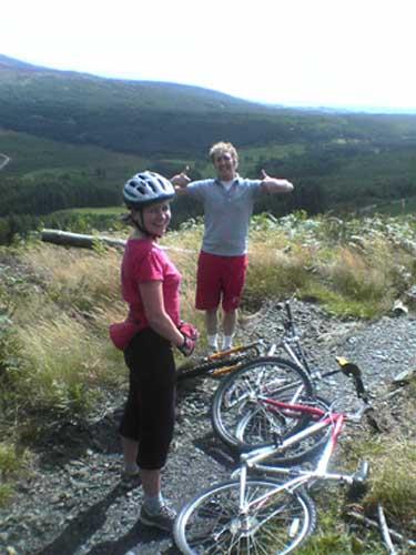 Two mountain bikers pause for a break on a rugged trail surrounded by greenery, with hills in the background. One person, wearing a helmet and a pink shirt, smiles at the camera, while the other gives a thumbs up. Two bicycles are on the ground nearby. Kirroughtree mountain bike trail.