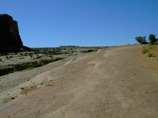 A rocky landscape under a clear blue sky, featuring a wide, flat path leading towards a distant hill. Sparse vegetation is visible along the edges, and a prominent rock formation is positioned to the left. Monitor &amp; Merrimac/Courthouse Pasture mountain bike trail.