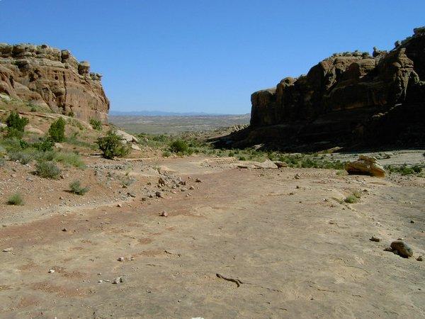 A wide view of a dry, rocky landscape featuring steep cliffs on either side, with sparse vegetation and a clear blue sky above. The terrain is mostly barren, with some small shrubs scattered throughout, leading into the distance where more hills and mountains are visible. Monitor &amp; Merrimac/Courthouse Pasture mountain bike trail.