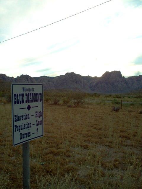A roadside sign welcoming visitors to Blue Diamond, featuring information about the town's high elevation, low population, and the presence of burros, set against a backdrop of mountains and an open desert landscape under an overcast sky. Blue Diamond mountain bike trail.