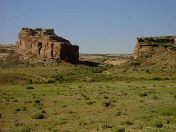 A desert landscape featuring two prominent rock formations with a grassy area in the foreground under a clear blue sky. The rock formations have varying shades of tan and brown, with some vegetation visible in the surrounding area. Monitor &amp; Merrimac/Courthouse Pasture mountain bike trail.