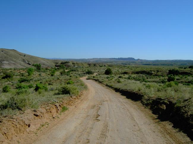 A winding dirt road stretches through a grassy landscape under a clear blue sky, surrounded by low hills and distant mountains. The scene captures the tranquility of a rural setting with sparse vegetation and rocky terrain. Monitor &amp; Merrimac/Courthouse Pasture mountain bike trail.