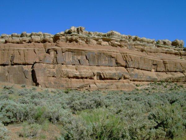 A rocky cliff with layered sandstone formations and sparse vegetation at the base, under a clear blue sky. Monitor &amp; Merrimac/Courthouse Pasture mountain bike trail.