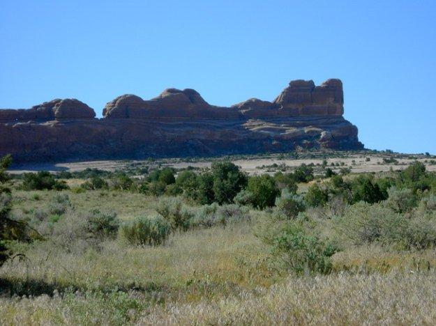 A panoramic view of a desert landscape featuring prominent rock formations in the background. The scene includes a clear blue sky and foreground vegetation with sparse shrubs and grasses. The rocky outcrop has a layered appearance, showcasing natural erosion patterns. Monitor &amp; Merrimac/Courthouse Pasture mountain bike trail.