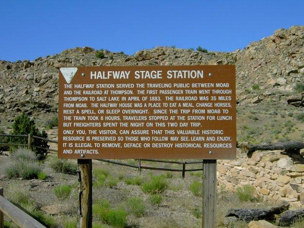 Sign at Halfway Stage Station, detailing its history as a stop for travelers between Moab and Thompson. The sign provides information on the station's role in the first passenger train journey through the area in 1883, serving as a resting place for lunch and overnight stays. It emphasizes the importance of preserving the site and warns against the removal or defacement of historical resources. Surrounding landscape features rocky terrain and sparse vegetation. Monitor &amp; Merrimac/Courthouse Pasture mountain bike trail.