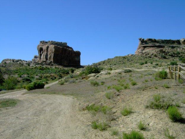 A scenic landscape featuring a dirt path winding through a grassy area, with rocky formations rising on either side under a clear blue sky. Monitor &amp; Merrimac/Courthouse Pasture mountain bike trail.