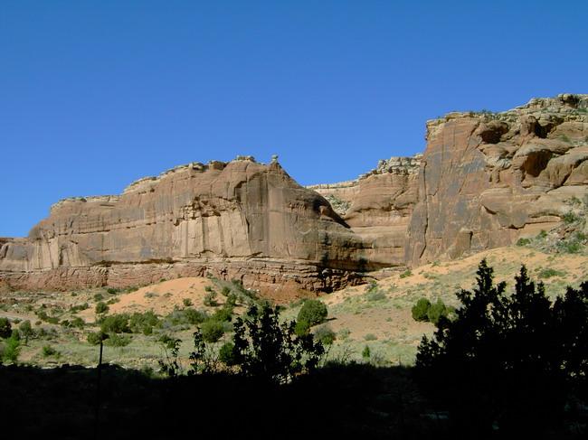 Rocky cliff formations rise against a clear blue sky, with patches of vegetation and sandy terrain visible below. Monitor &amp; Merrimac/Courthouse Pasture mountain bike trail.