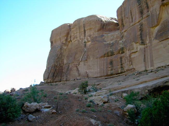 A view of a rugged rock formation towering against a clear blue sky, with a dirt path leading towards the base and sparse vegetation visible in the foreground. Monitor &amp; Merrimac/Courthouse Pasture mountain bike trail.