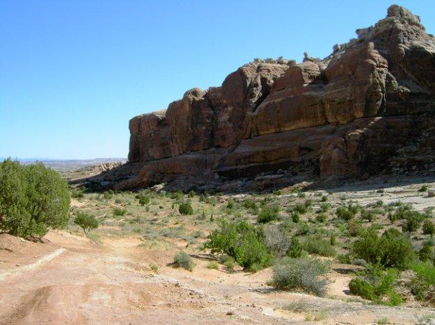 A rugged landscape featuring a desert scene with a dirt path leading toward large rock formations on the right. The sky is clear and blue, with sparse vegetation including low shrubs scattered across the arid terrain. Monitor &amp; Merrimac/Courthouse Pasture mountain bike trail.