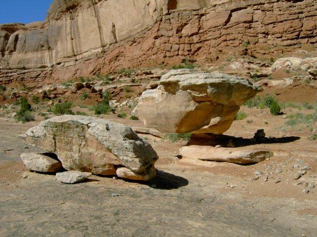 Two large boulders sit on a rocky desert landscape, with a backdrop of red cliff formations and sparse vegetation. The boulders are irregularly shaped, with one appearing to balance on top of the other. The scene captures the natural beauty of the area, highlighting the contrasting colors of the rocks and the arid environment. Monitor &amp; Merrimac/Courthouse Pasture mountain bike trail.