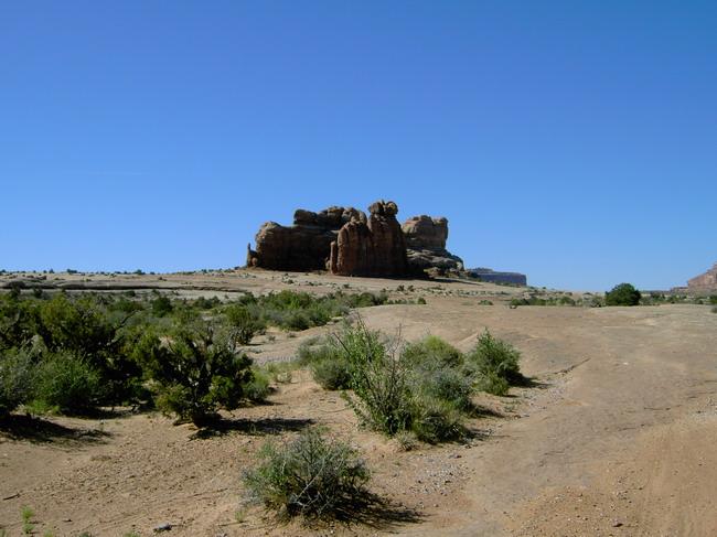 A rocky formation rises prominently against a clear blue sky, surrounded by arid land and sparse vegetation. The landscape features a dry, sandy path leading towards the rock structure, with scattered bushes creating a natural frame in the foreground. Monitor &amp; Merrimac/Courthouse Pasture mountain bike trail.